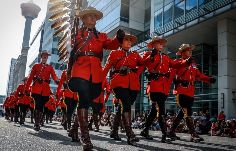 members of the RCMP in their fancy red dress uniforms march in a parade formation on a downtown Calgary street with the Calgary tower visible behind them.