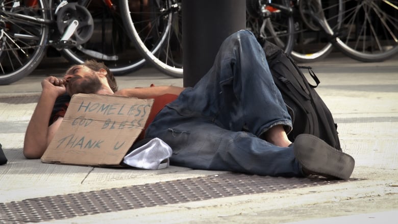 A man suffering from homelessness is lying down on the sidewalk. His sign reads, "Homeless. God Bless. Thank you."