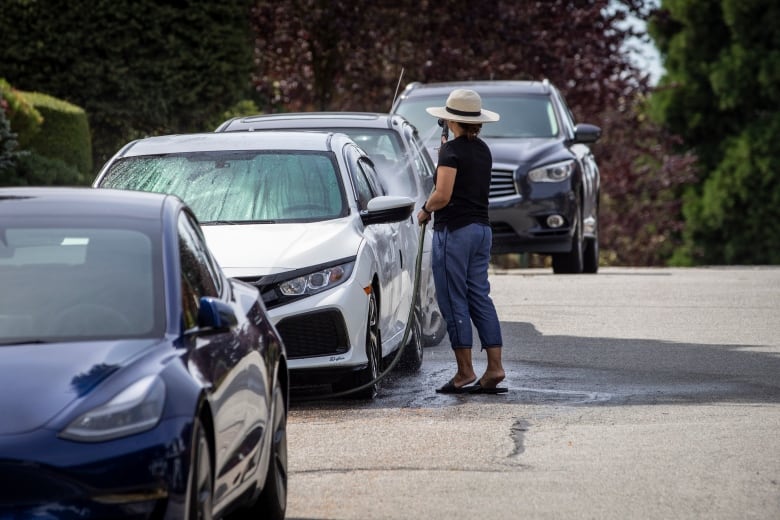 A woman in a black t-shirt, jeans and a hat is pictured watering her white sedan, among several cars lining a sidewalk. 