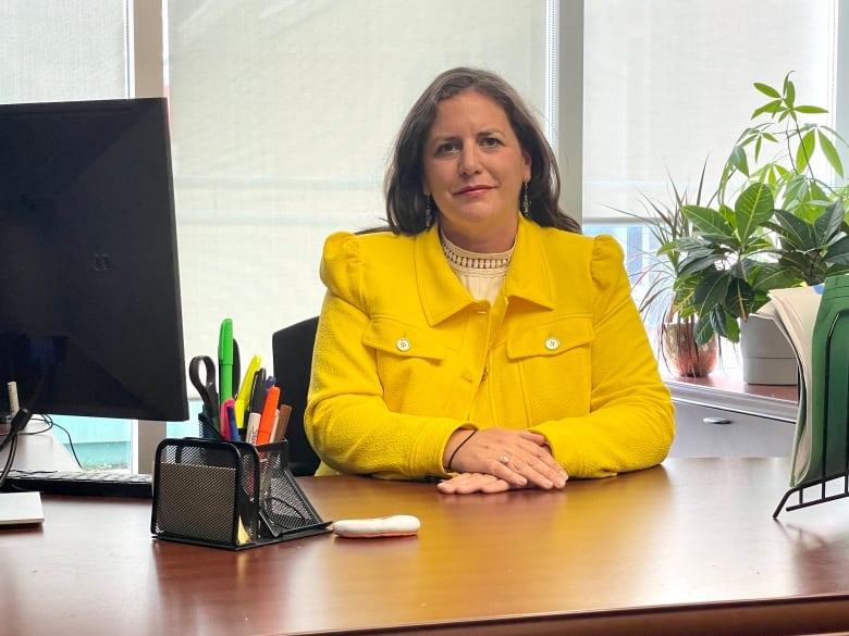 A woman with long brown hair and a yellow blazer sits at her desk. 