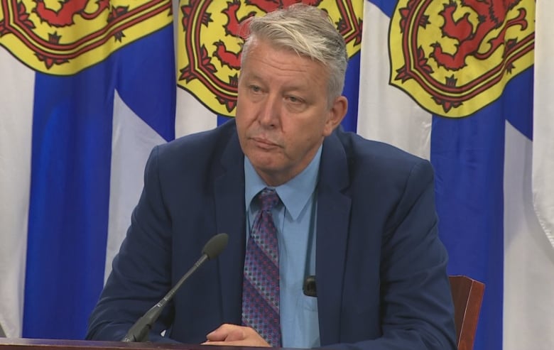 A man in a suit sits in front of a Nova Scotia flag. 