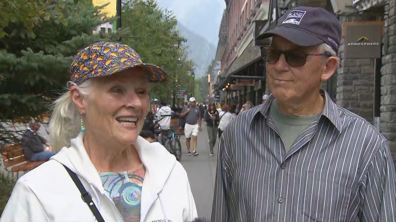 A women in a white hoodie and patterend baseball cap and a man in a striped button up shirt in a navy hat stand in Banff.
