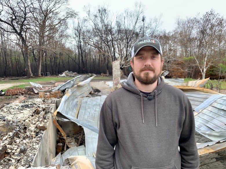 A man in a ball cap stands in front of a burned out house. 