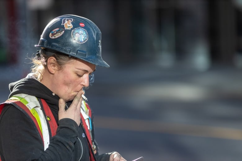 A construction worker looks at their phone while taking a drag from their cigarette.