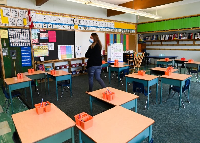 A female teacher walks past empty desks in a classroom. 