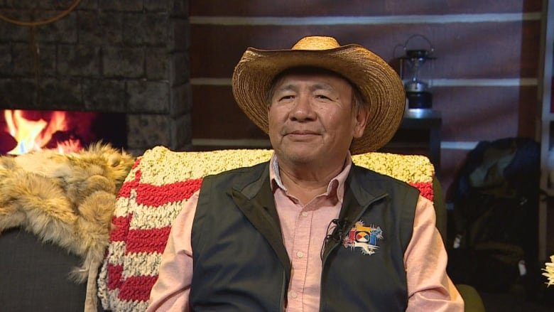 A man can be seen smiling, he is wearing a hat, a red shirt, a black jacket while sitting on a couch. 