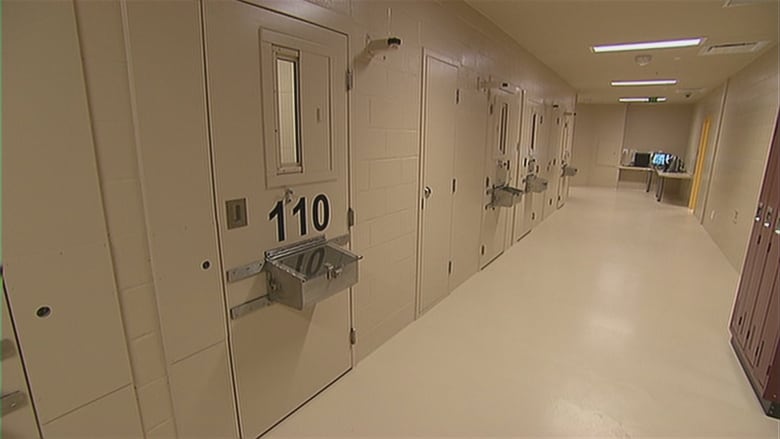 A long hallway in a provincial jail is pictured. There are several doors to cells with numbers printed on them, and silver lockboxes outside each door.