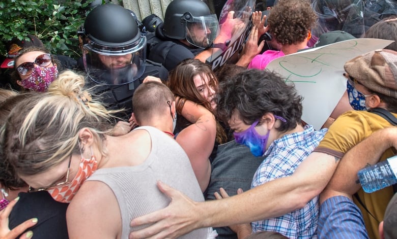 A crowd of people wrap arms around each other as they stand against police in black helmets carrying riot gear.