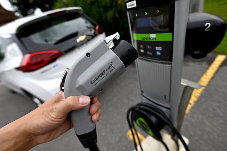 A hand holds a grey EV charger at a gas station. 