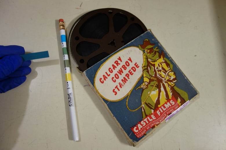 A hand holds a blue strip up beside a Calgary Stampede film reel. 