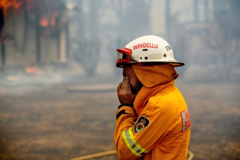 A firefighter in a bright orange jacket and a white helmet rubs his eyes with his hands as smoke and haze hover in the air around him. 