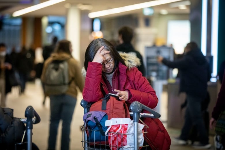 A frustrated passenger in a red parka rests their head in their hands and leans on a baggage cart at Vancouver International Airport.