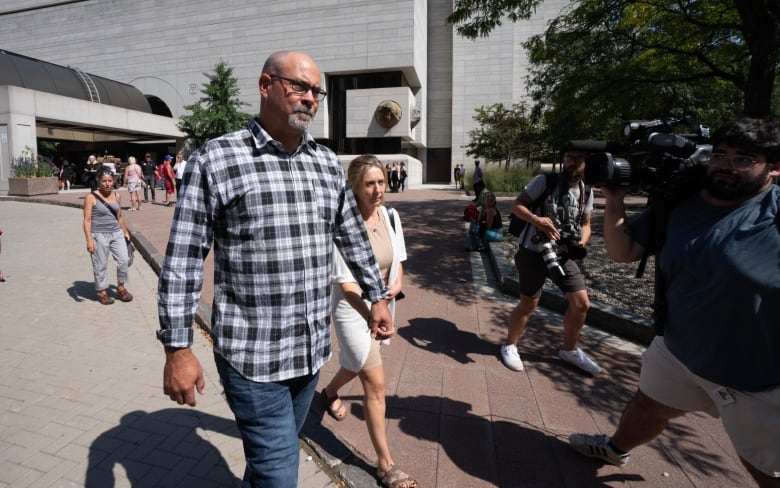 Two people walk away from a courthouse in summer as videographers aim cameras as them.
