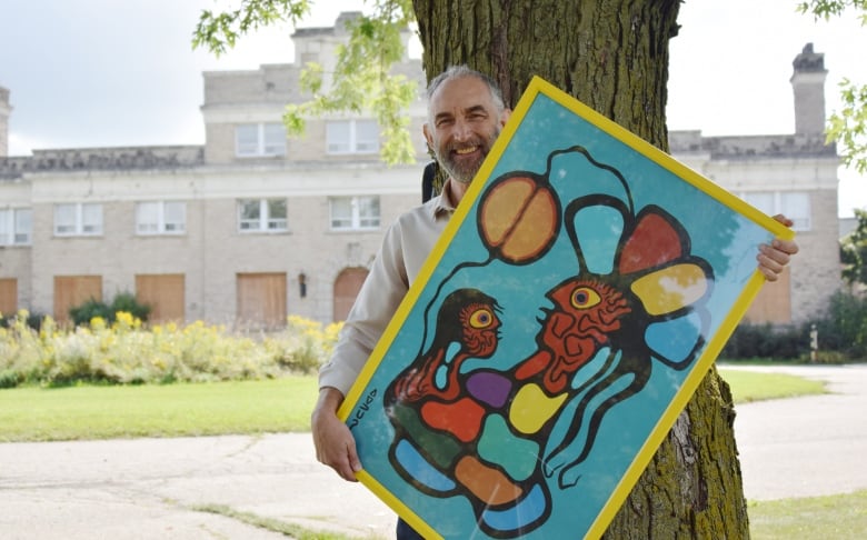 Man holds up colourful Indigenous painting standing in front of an old building - the former jail