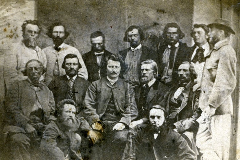 Black and white photo of a group of 15 men from the 1870s. Most are standing, surrounding one man seated in the centre.