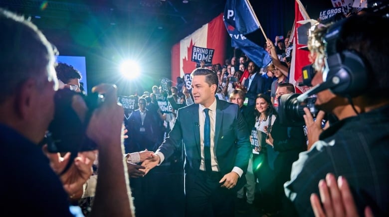 A man in a suit shakes hands with a crowd as he enters a convention hall.