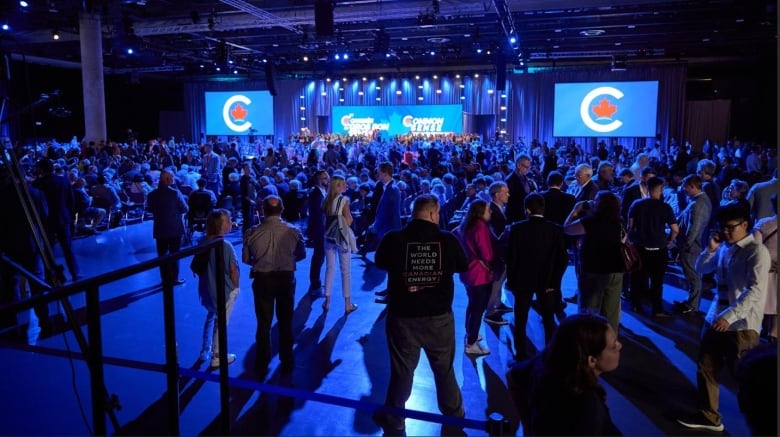 A crowd fills a convention hall marked with Conservative party logos.