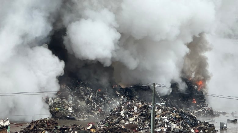 Flames leap out of a large pile of scrap metal, smoke billows