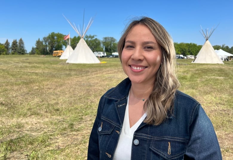 A woman in a denim jacket smiles at the camera. Behind her, several teepees are set up. The dry grass is a combination of green and yellow, with a cloudless sky above.