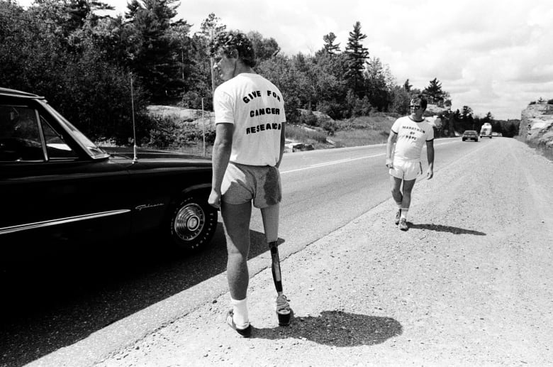 a black and white photo of Terry Fox wearing a t-shirt that says Give for Cancer Research and Bill Vigars is in the background. There is dark car parked beside them. 