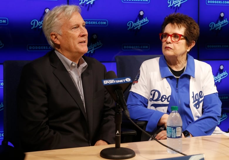 A white haired man and a dark haired woman with red glasses sit together at a table with a screen of sports logos in back of them and a microphone on the table
