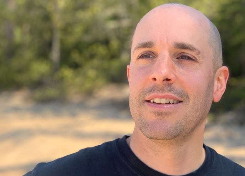 A man with a shaved head looks off camera while standing in a sunny wooded area. 