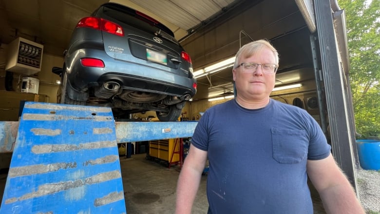 An older man with short grey hair and glasses faces the camera with a neutral facial expression. He is wearing a dark blue t-shirt. Behind him is a dark blue SUV lifted up on a garage hoist.