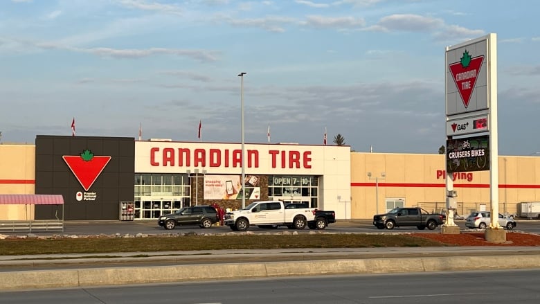A storefront shows the Canadian Tire logo with large sliding automatic doors as the entrance. The sky is dark and cloudy and three pickup trucks and an SUV are stopped in the parking lot in front of the doors.