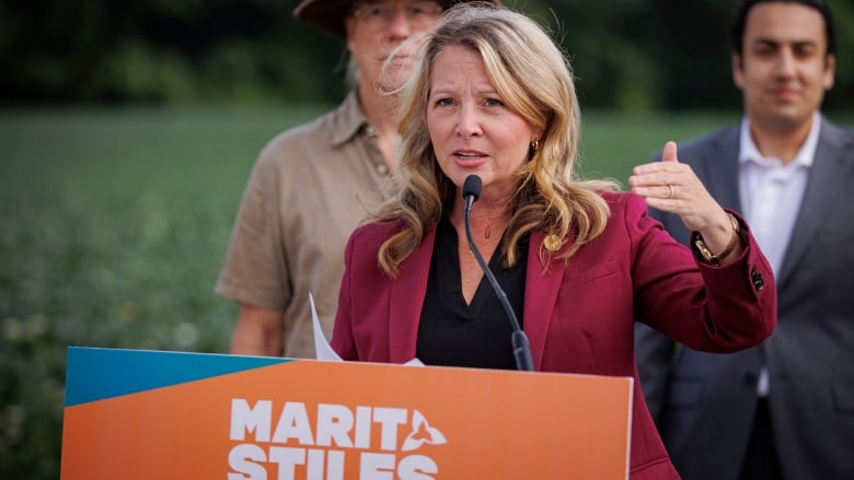 Marit Stiles stands at a podium outdoors, with a field in the background. 