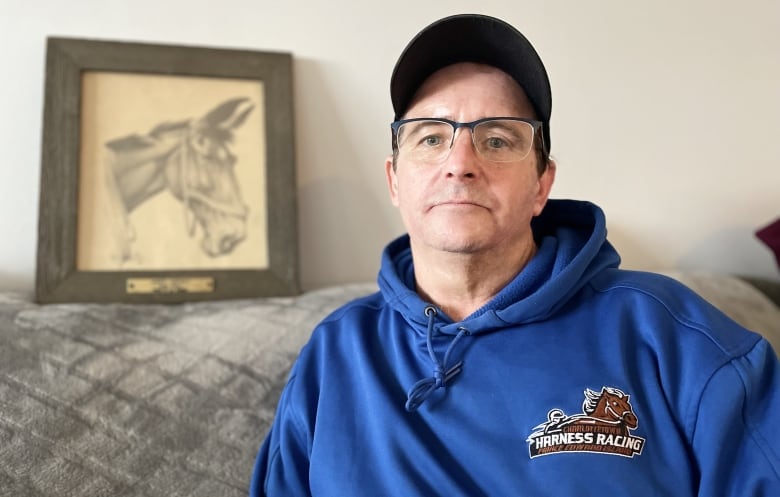 A man with glasses and a baseball cap wears a blue hoodie with a harness racing logo. A drawing of a horse its in the background.
