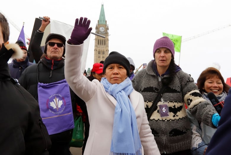 A crowd of people in winter clothes standing in front of Ottawa's Parliament building raise their hands in the air. The Parliament building's steeple with the clock and green tower are in the background.