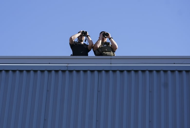 Two police officers on a rooftop look through binoculars.