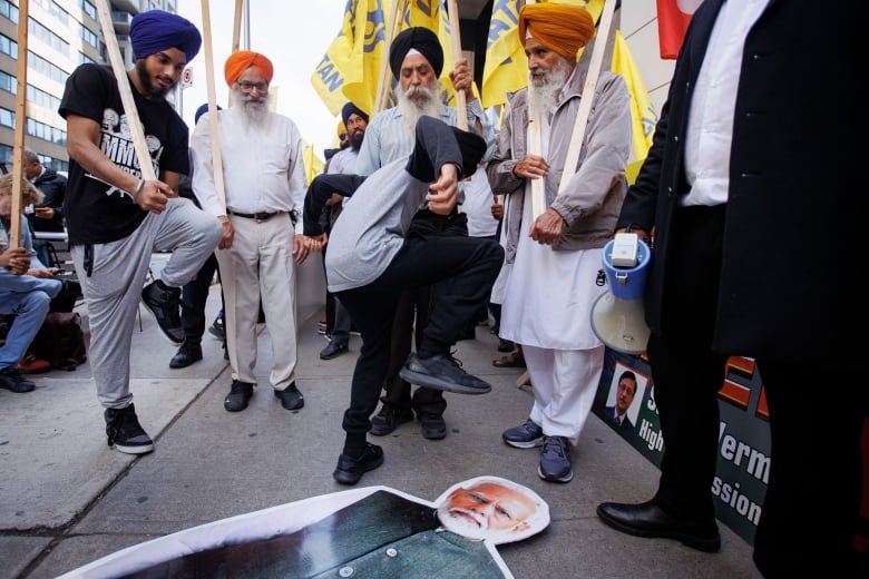 A group of men in turbans hold signs and stomp on a cutout of Indian Prime Minister Narendra Modi on the ground. 