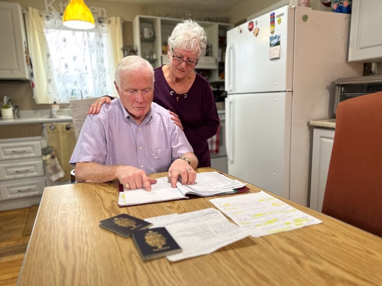 An elderly white man and woman look at documents on their wooden kitchen table. 