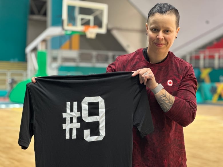 A woman holds up a black shirt with the number nine emblazoned on it. A basketball court is in the background.