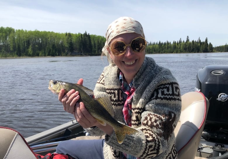 A woman wearing sunglasses, a head scarf and a Cowichan sweater sits in a boat on the water while smiling and holding a fish. 