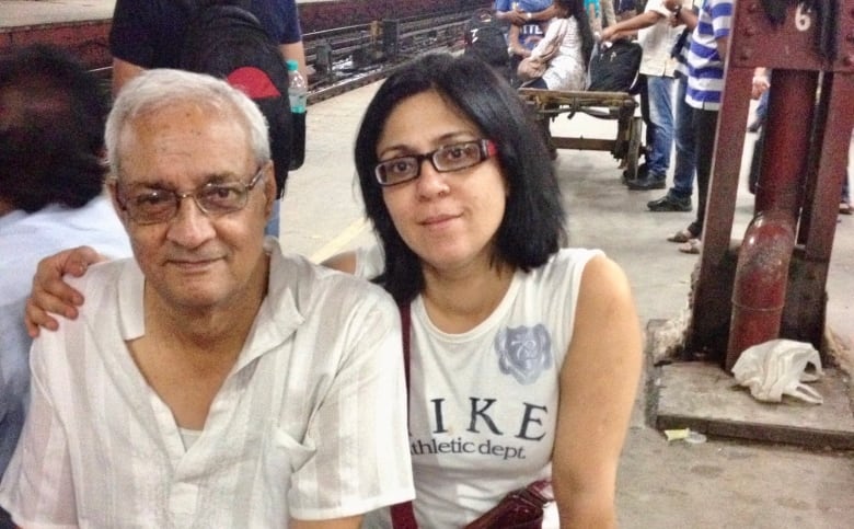 An aged south Asian man and a south Asian woman sit on a bench on a busy train platform. The woman has her right arm around the man. 