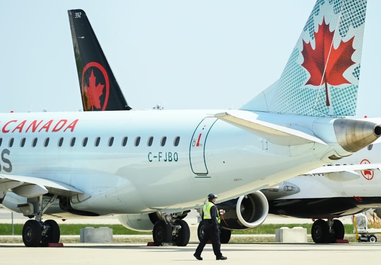 Two Air Canada jetliners sit on a runaway. A man walks in front of them. 