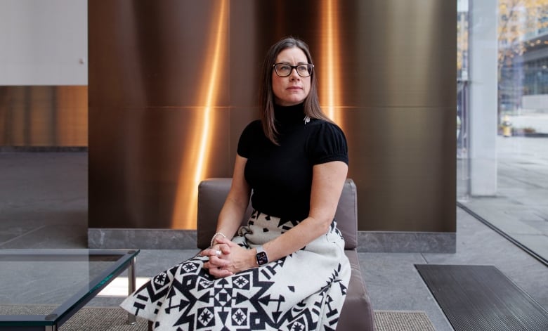 A waman with long brown hair and glasses sits in a hotel lobby with 2 golden beams of light in the background. She is wearing a black sweater and white skirt with an Indigenous themed print on it. 