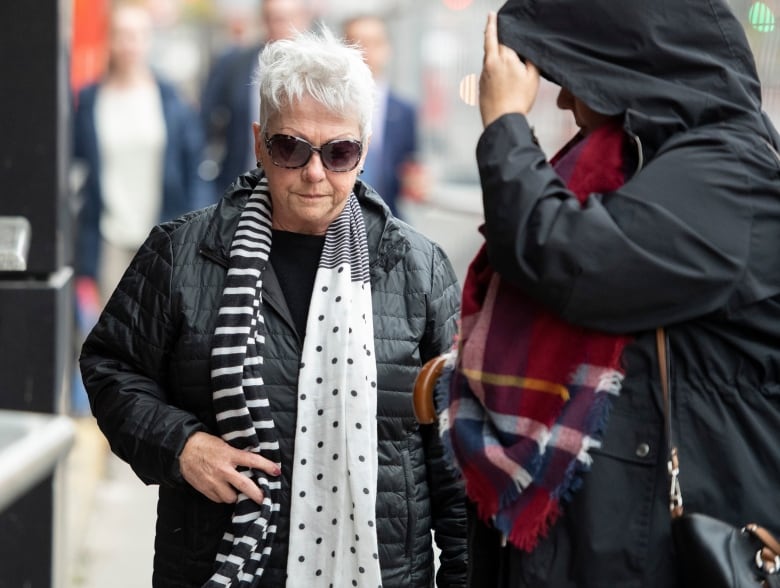 An older woman in dark glasses walks down a street.