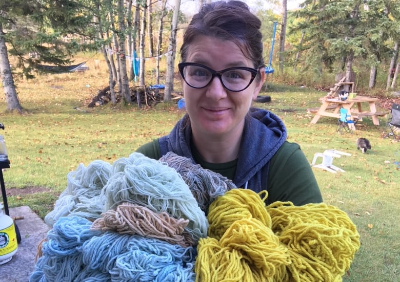 A woman in glasses holds skeins of yarn in several different colours. 
