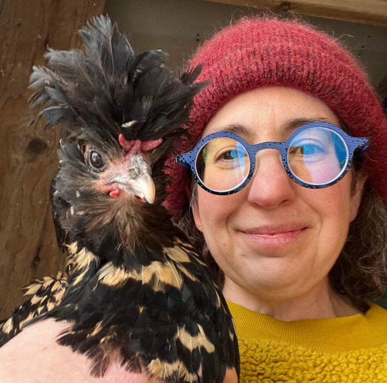A woman wearing a knitted hat and sweater takes a selfies while holding a chicken. 