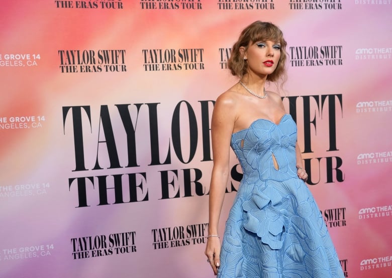 A blonde woman with a bob hair cut wears a blue glown while posing in front of a pink billboard that says Taylor Swift.