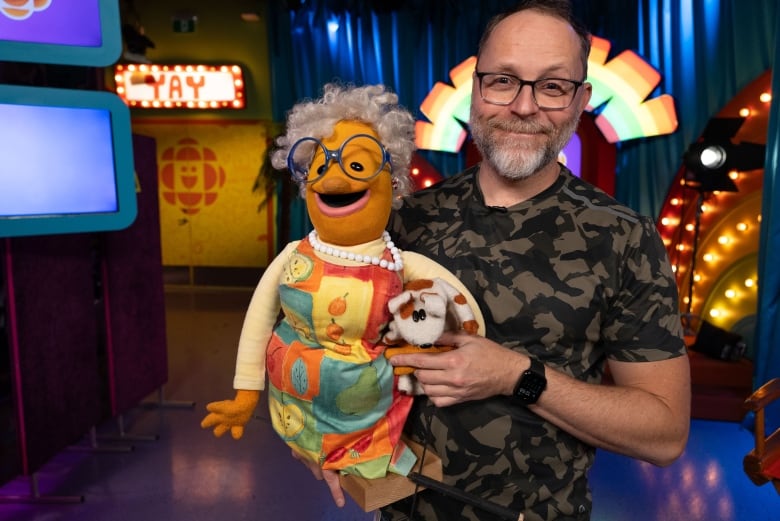 A man with glasses holds two puppets: one grandmother with a colourful dress, big glasses and white pearls and a small Jack Russell-like dog