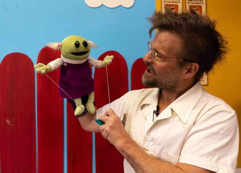 A man in a white shirt and glasses holds a puppet. The puppet has a green pea for a head, black beady eyes, pig tails and purple dress.