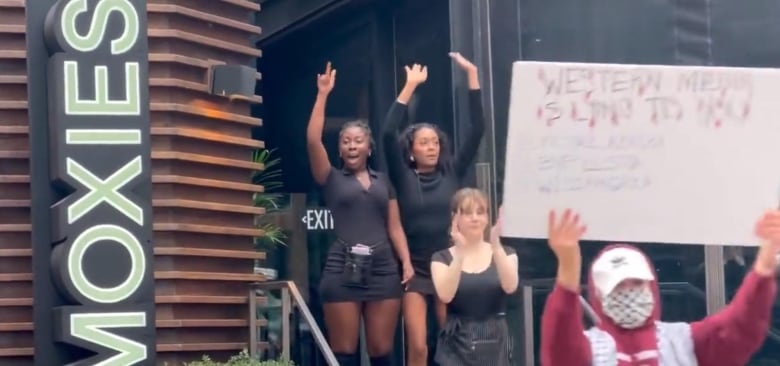 People standing at the door of Moxies restaurant in black uniforms, with arms raised. In the foreground, a protester carries a sign.