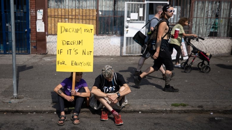 A woman in sandals and a man in red shoes sit on the curb of Vancouver's East Hastings street. The woman has a tatoo on her right arm and is holding a yellow cardboard sign attached to a piece of wood that reads, "Decrim isn't decrim if it's not for everyone." The man is wearing a camo baseball cap and has his head bowed. Three people are walking in the background.