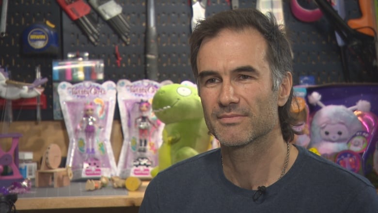 A man sits in front of a desk filled with toys including a green dinosaur. 