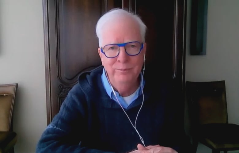 Man with white hair, blue glasses sits at desk wearing blue sweater.