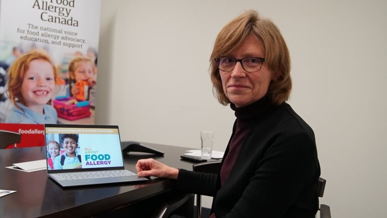 Sitting in an indoor meeting room, a woman in a black blazer and glasses turns to smile at the camera as she scrolls through a laptop showing slides about food allergies. 